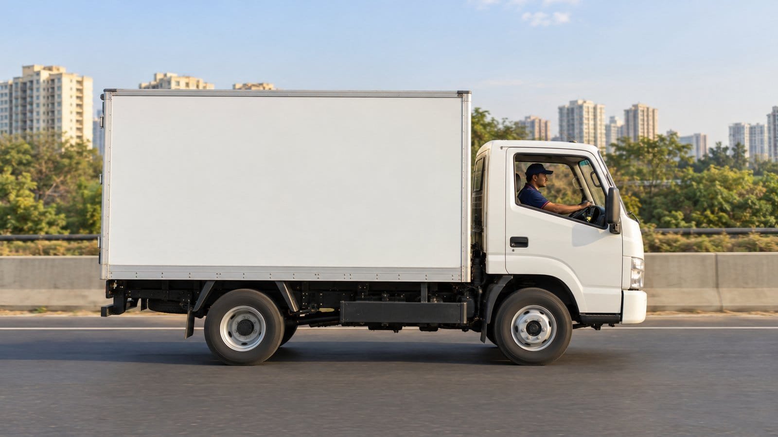 A loaded moving truck on a multi-lane highway with a flyover and a distant city skyline, route markers pointing in two directions